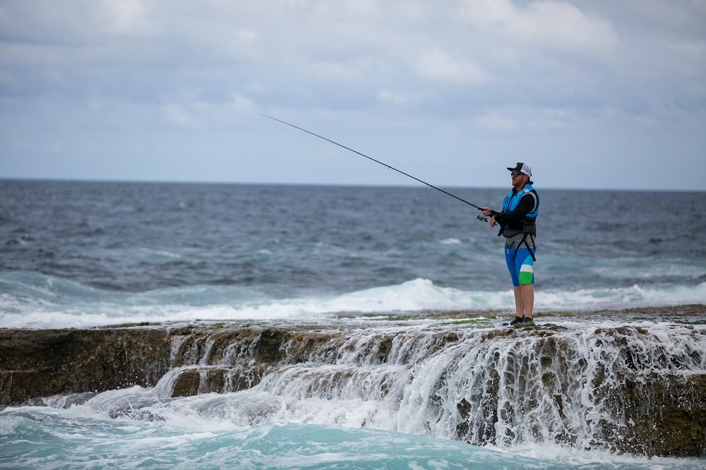 A man wearing a life jacket stands fishing on a rock shelf with shallow water running back into the ocean.